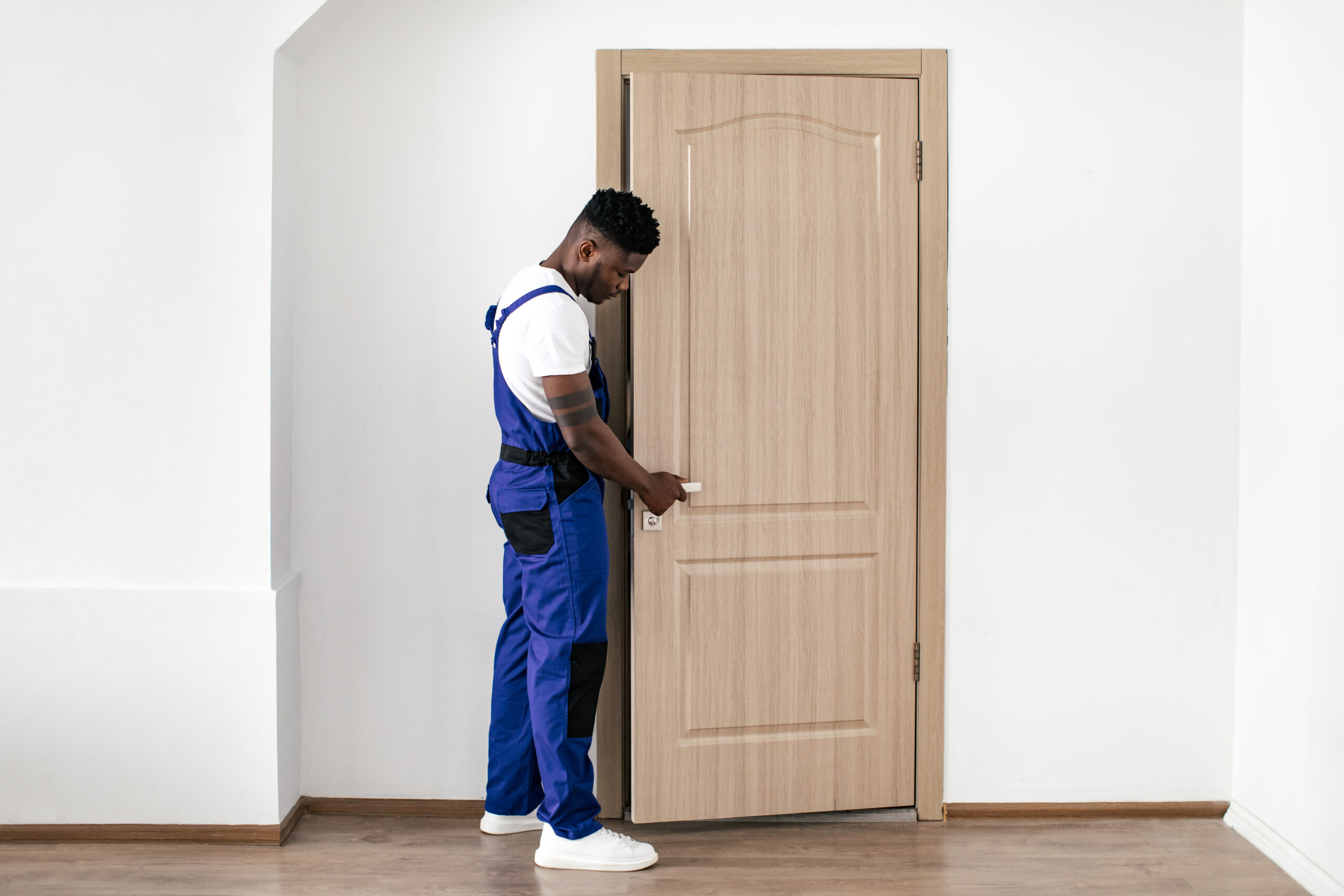 Repair Service. Black Repairman Installing New Entry Door, Fixing Lock And Handle Working Wearing Blue Coverall Uniform In Flat Indoor. Male Renovation Concept. Full Length Shot