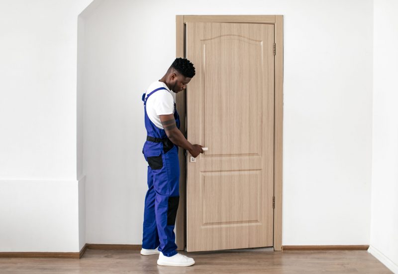 Repair Service. Black Repairman Installing New Entry Door, Fixing Lock And Handle Working Wearing Blue Coverall Uniform In Flat Indoor. Male Renovation Concept. Full Length Shot