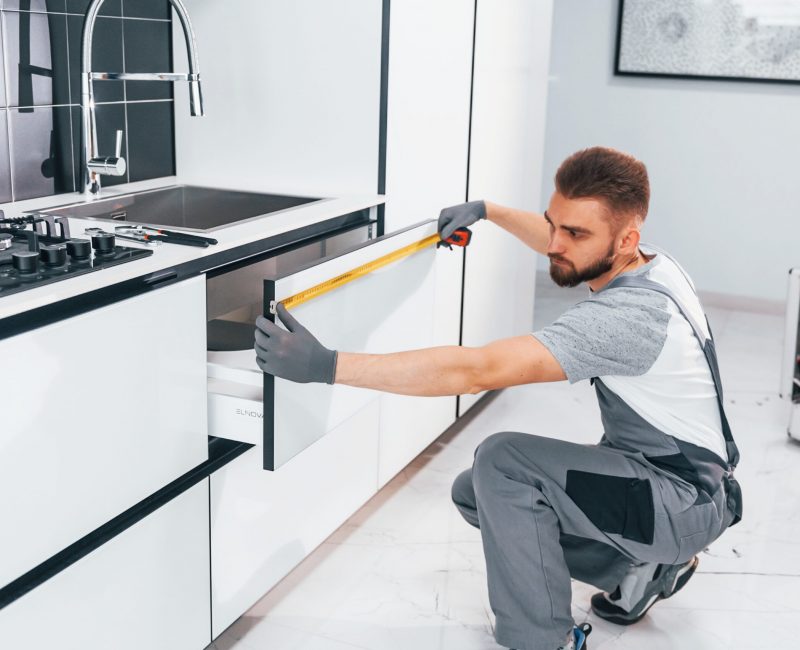 Young professional plumber in grey uniform measuring table by using meter on the kitchen.